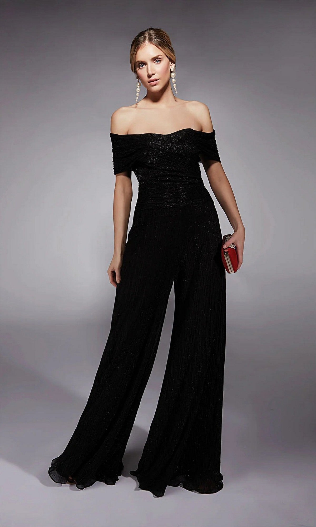 A woman in the Alyce 27768 Long Formal Jumpsuit in black with wide legs, paired with a red clutch and pearl earrings, stands against a plain gray background.